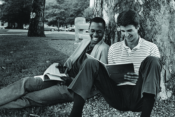 Black and white photograph of two students laughing and studying under a tree