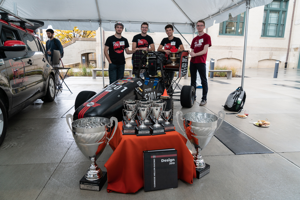 Student racing team posing with their car as well as numerous trophies they have received