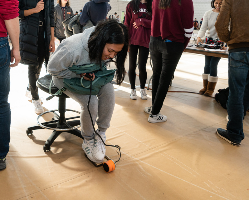 A student sitting on a rolling chair and pressing on a level on the floor with her shoes